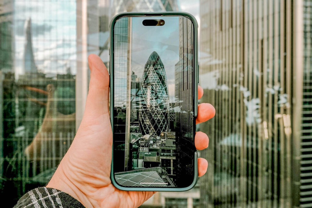 Photo of a hand holding a mobile phone showing a picture of the Gherkin in London in front of the Gherkin in London